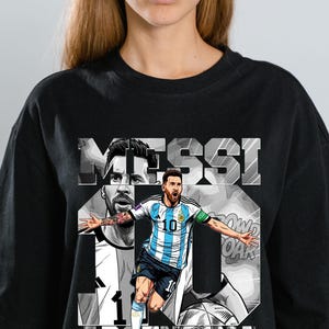 Può includere: T-shirt nera con una grafica di un calciatore con la maglia dell'Argentina, con il nome "MESSI" e il numero "10" a lettere grandi. La parola "ARGENTINA" è stampata in basso.
