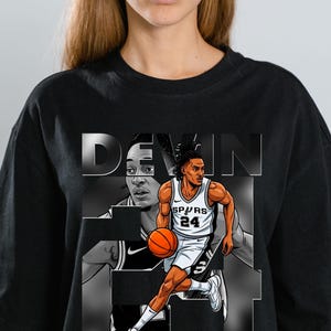 Pode incluir: Camiseta preta com um gráfico do jogador de basquete Devin Vassell driblando uma bola de basquete laranja. O design inclui o nome e o número da camisa do jogador, com uma imagem em tons de cinza do jogador no fundo.