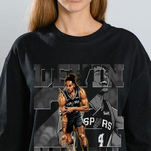 Pode incluir: Uma t-shirt preta de manga curta com uma estampa gráfica do jogador de basquete Devin Vassell com o uniforme do San Antonio Spurs. O design inclui o texto "DEVIN" e "VASSELL" em letras grandes, juntamente com um número 24 grande.