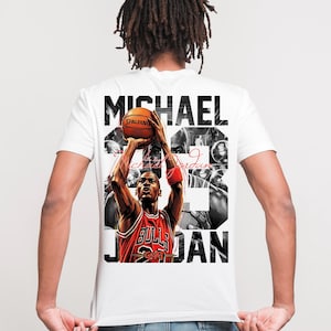 May include: White t-shirt featuring a graphic of Michael Jordan in a red and black Chicago Bulls jersey, shooting a basketball. The shirt has the name "Michael Jordan" in large black letters.