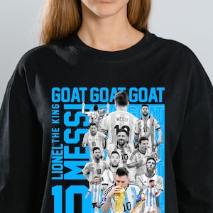 Peut inclure: T-shirt noir avec un motif bleu représentant Lionel Messi en maillot argentin. Le design inclut les mots "GOAT" et "MESSI 10". Le t-shirt est de style col rond.