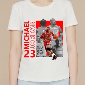 May include: White t-shirt featuring a collage of Michael Jordan images in red and grayscale. The design includes the text "MICHAEL JORDAN 23" in red vertical lettering. The images show Jordan in his Bulls uniform.