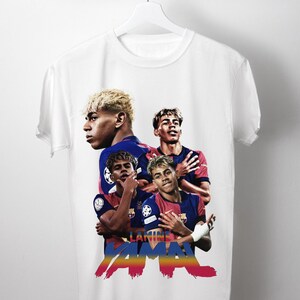 May include: White t-shirt with a design featuring a soccer player in a blue and red uniform. The design includes the player's image and the name "LAMINE YAMAL" in a colourful, stylised font.