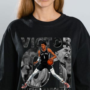 Puede incluir: Camiseta negra con un gráfico de un jugador de baloncesto con el uniforme de los Spurs, sosteniendo un balón de baloncesto. El diseño incluye el nombre "VICTOR WEMBANYAMA" en negrita. La camiseta tiene cuello redondo.