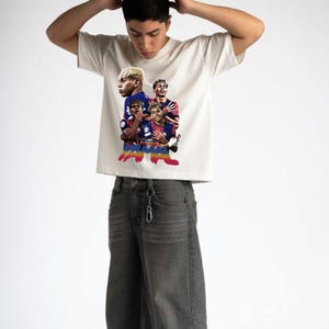 May include: Cream-coloured t-shirt featuring a graphic of a football team. The graphic includes images of the players in their team uniforms. The person in the image is wearing grey jeans.