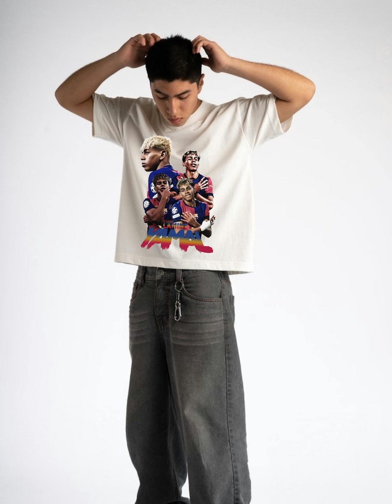 May include: Cream-coloured t-shirt featuring a graphic of a football team. The graphic includes images of the players in their team uniforms. The person in the image is wearing grey jeans.