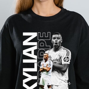 Op de afbeelding: Zwart T-shirt met ronde hals en een zwart-wit afbeelding van een voetballer. De naam "KYLIAN MBAPPE" is verticaal in witte blokletters gedrukt. Sportief thema.