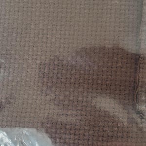 Puede incluir: Primer plano de una tela marrón tejida con una superficie texturizada. La tela parece estar tejida apretadamente, con un patrón consistente de pequeños cuadrados. El material está encerrado en una bolsa de plástico transparente.