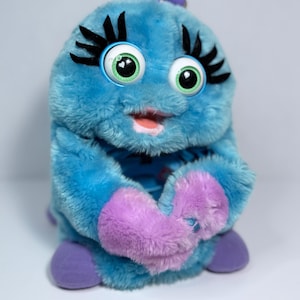May include: A plush toy in shades of blue and purple, featuring large, expressive eyes with black eyelashes and a small purple antenna. The toy has a smiling mouth and soft, fluffy texture, with purple accents on the hands and feet.