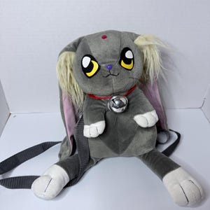 Op de afbeelding: Een grijze pluche rugzak in de vorm van een konijn met lange oren, gele en zwarte ogen, een kleine paarse neus en een rode halsband met een zilveren bel. De rugzak heeft witte pootjes, crèmekleurige vachtaccenten en verstelbare grijze schouderbanden.