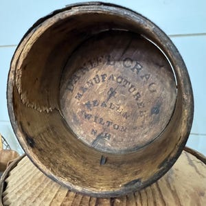 May include: An antique wooden barrel with visible wear and tear. The interior base features the text "DANIEL CRAG MANUFACTURER & DEALER WILTON N.H." in a circular arrangement. The barrel's aged appearance suggests it is vintage.