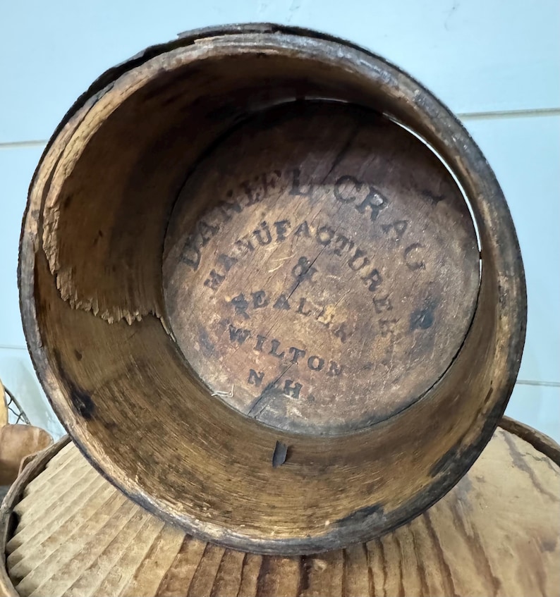 May include: An antique wooden barrel with visible wear and tear. The interior base features the text "DANIEL CRAG MANUFACTURER & DEALER WILTON N.H." in a circular arrangement. The barrel's aged appearance suggests it is vintage.