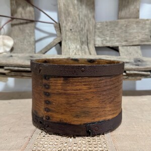 May include: A cylindrical wooden container with a dark brown metal band around the top and bottom. The wood has a warm, natural tone, and the metal bands are riveted to the wood. The container is likely vintage or antique.