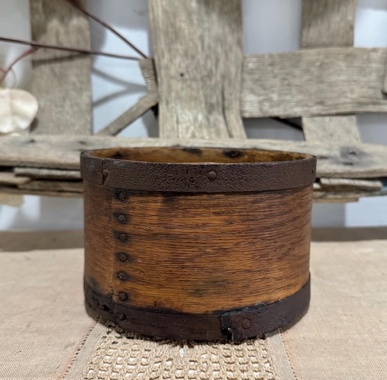 May include: A cylindrical wooden container with a dark brown metal band around the top and bottom. The wood has a warm, natural tone, and the metal bands are riveted to the wood. The container is likely vintage or antique.