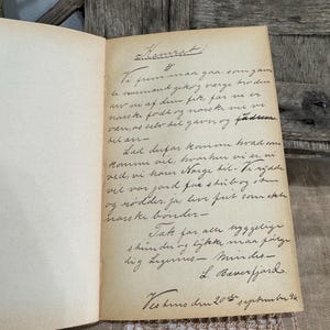 May include: An open vintage book with handwritten text in black ink on aged, cream-colored pages. The text is in a cursive script, with the title "Kamrat" at the top. The book is resting on a wooden surface.