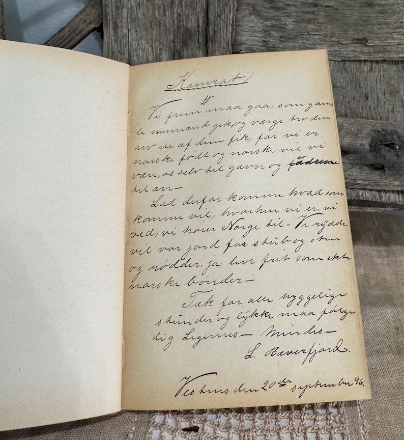 May include: An open vintage book with handwritten text in black ink on aged, cream-colored pages. The text is in a cursive script, with the title "Kamrat" at the top. The book is resting on a wooden surface.
