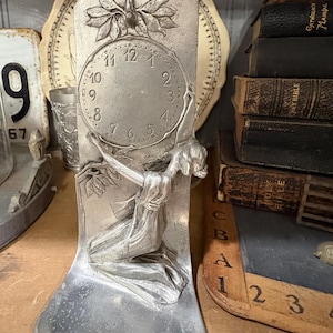 May include: A silver-toned Art Nouveau clock featuring a kneeling female figure with outstretched arms. The clock face has Roman numerals and is adorned with floral details. A clear glass tube sits atop the clock. The clock is displayed with vintage books and other antique items.