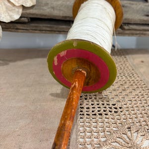 May include: An antique wooden thread spool with white thread. The spool has a wooden handle and a circular top with green and pink accents. The spool is resting on a beige crocheted surface.