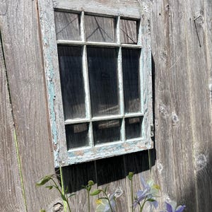 May include: A rectangular wooden window frame with six glass panes, featuring weathered, chipped light blue paint over a natural wood finish. The frame is mounted vertically and displays a grid structure with thin muntins separating the glass sections.