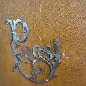 May include: A decorative metal sign with the word "Poes" in an ornate, vintage font. The sign is silver-colored with a distressed finish, and is set against a textured, tan background.