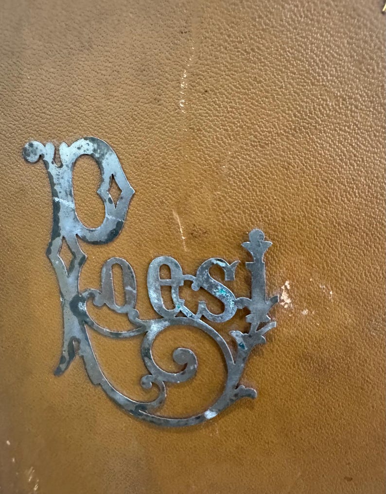 May include: A decorative metal sign with the word "Poes" in an ornate, vintage font. The sign is silver-colored with a distressed finish, and is set against a textured, tan background.