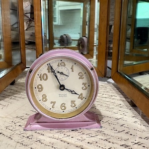 May include: A round, light pink metal Westclox Baby Ben alarm clock with a cream-colored face, black numerals, and black hands. The clock features a smaller sub-dial at the top and the text Westclox Baby Ben printed on the center of the face.