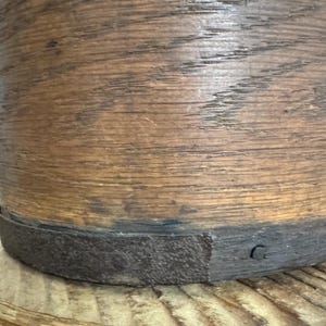 May include: A cylindrical wooden container with a dark metal band around the base. The wood has a natural, aged appearance with visible grain and a warm brown color. The metal band is dark gray.