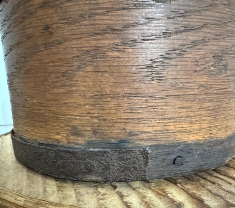 May include: A cylindrical wooden container with a dark metal band around the base. The wood has a natural, aged appearance with visible grain and a warm brown color. The metal band is dark gray.