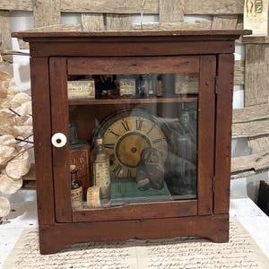 May include: A brown wooden cabinet with a glass door and a white knob. Inside are various items including a round clock with Roman numerals, glass bottles with labels, a dark metal padlock, a green book, and small boxes labeled Sand and Fourths.