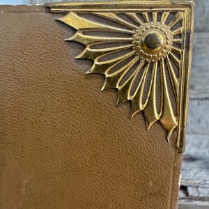 May include: Close-up of a vintage book corner with a gold-colored metal embellishment. The corner protector features a sunburst design. The book's cover is a textured, tan color.