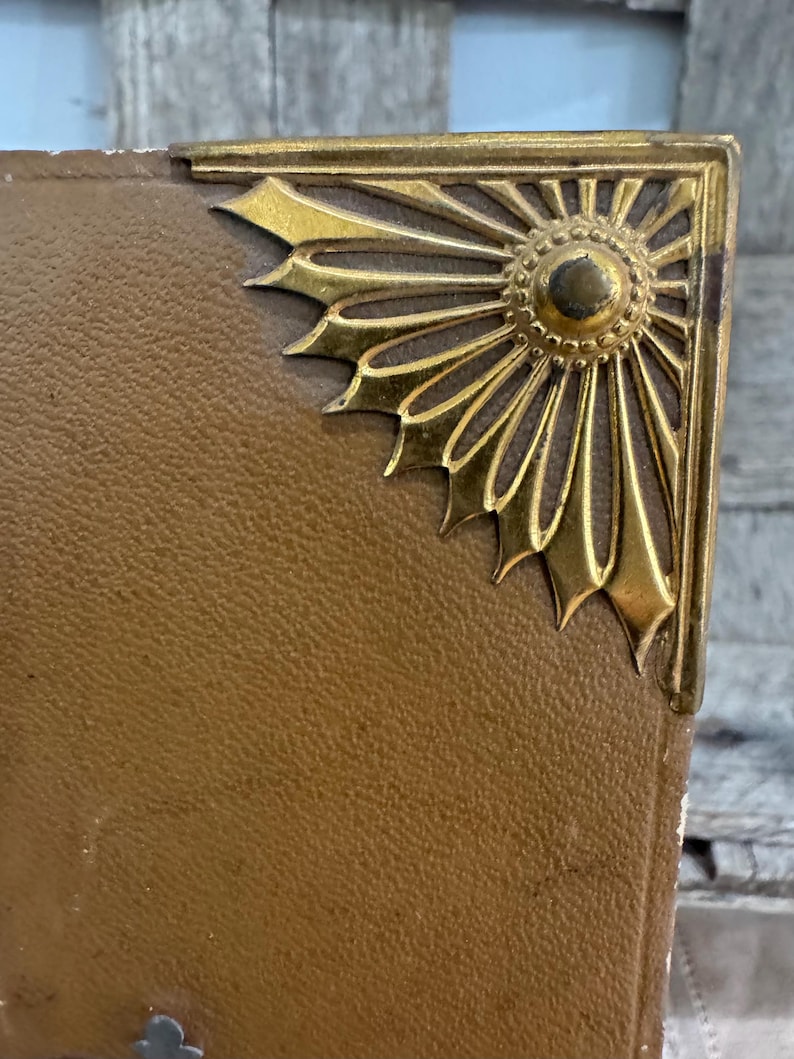 May include: Close-up of a vintage book corner with a gold-colored metal embellishment. The corner protector features a sunburst design. The book's cover is a textured, tan color.