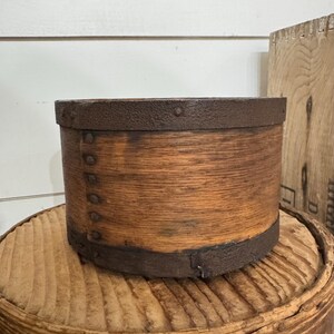 May include: A cylindrical wooden container with a rustic, aged appearance. The container is made of light brown wood with dark brown metal bands around the top and bottom. Small metal rivets secure the bands. The container sits on a wooden surface.