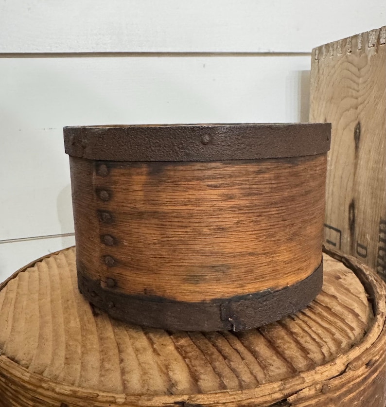 May include: A cylindrical wooden container with a rustic, aged appearance. The container is made of light brown wood with dark brown metal bands around the top and bottom. Small metal rivets secure the bands. The container sits on a wooden surface.