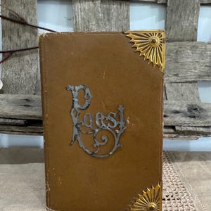 May include: A vintage book with a brown cover and gold corner accents. The cover features the word "Poest" in an ornate, silver-colored font. The book is slightly worn, suggesting its age and history.