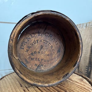 May include: A weathered, cylindrical wooden container with visible grain and a dark brown hue. The interior base features the stamped text "DANIEL CRAG MANUFACTURER DEALER WILTON N.H."