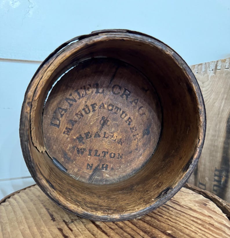 May include: A weathered, cylindrical wooden container with visible grain and a dark brown hue. The interior base features the stamped text "DANIEL CRAG MANUFACTURER DEALER WILTON N.H."