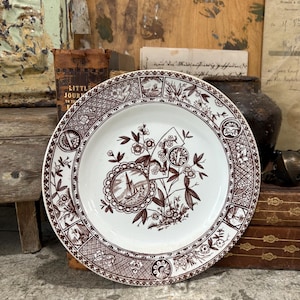 May include: A white and brown antique plate with a detailed floral and nautical design. The plate features a central medallion with a sailboat and is surrounded by intricate patterns and borders. The plate is a vintage piece.