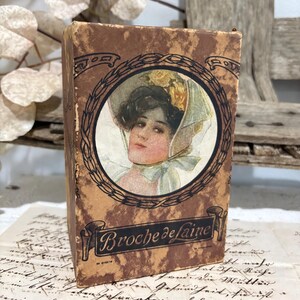 May include: A rectangular cardboard box with a mottled brown finish featuring a central circular illustration of a person wearing a bonnet with yellow flowers. The text Broche de laine is printed in black script on a banner at the bottom of the box.