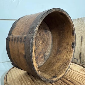 May include: A vintage wooden barrel with metal bands. The barrel is made of light brown wood and has a circular shape. The metal bands are dark brown and encircle the barrel. The interior of the barrel has visible text. The barrel is sitting on a wooden surface.