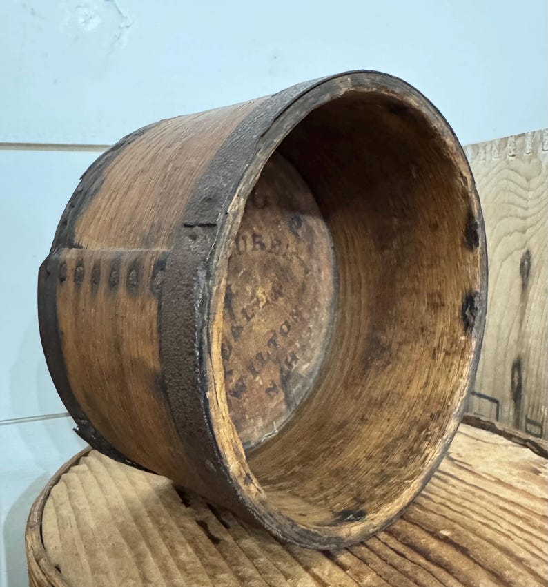 May include: A vintage wooden barrel with metal bands. The barrel is made of light brown wood and has a circular shape. The metal bands are dark brown and encircle the barrel. The interior of the barrel has visible text. The barrel is sitting on a wooden surface.