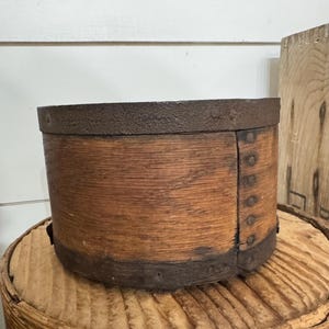 May include: A round, wooden container with a dark metal rim and accents. The container has a rustic, aged appearance, with visible wood grain and metal rivets. The container is sitting on a round, wooden surface.
