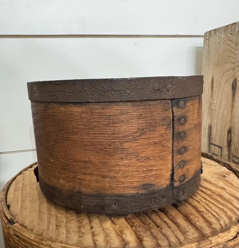 May include: A round, wooden container with a dark metal rim and accents. The container has a rustic, aged appearance, with visible wood grain and metal rivets. The container is sitting on a round, wooden surface.