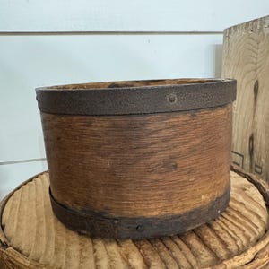 May include: A cylindrical wooden container with dark metal bands around the top and bottom. The wood has a natural, aged appearance. The container is sitting on a woven basket, suggesting a rustic or farmhouse aesthetic.