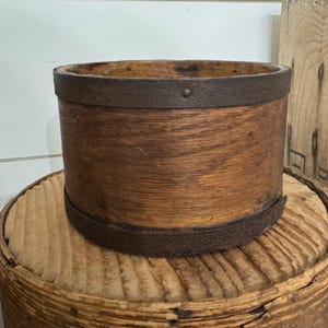 May include: A cylindrical wooden container with a rusty metal band around the top and bottom. The wood has a warm, natural tone, and the metal bands add a rustic touch. The container sits on a round, wooden surface.
