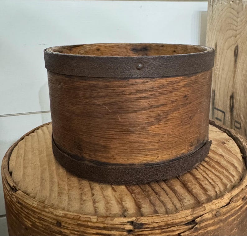 May include: A cylindrical wooden container with a rusty metal band around the top and bottom. The wood has a warm, natural tone, and the metal bands add a rustic touch. The container sits on a round, wooden surface.
