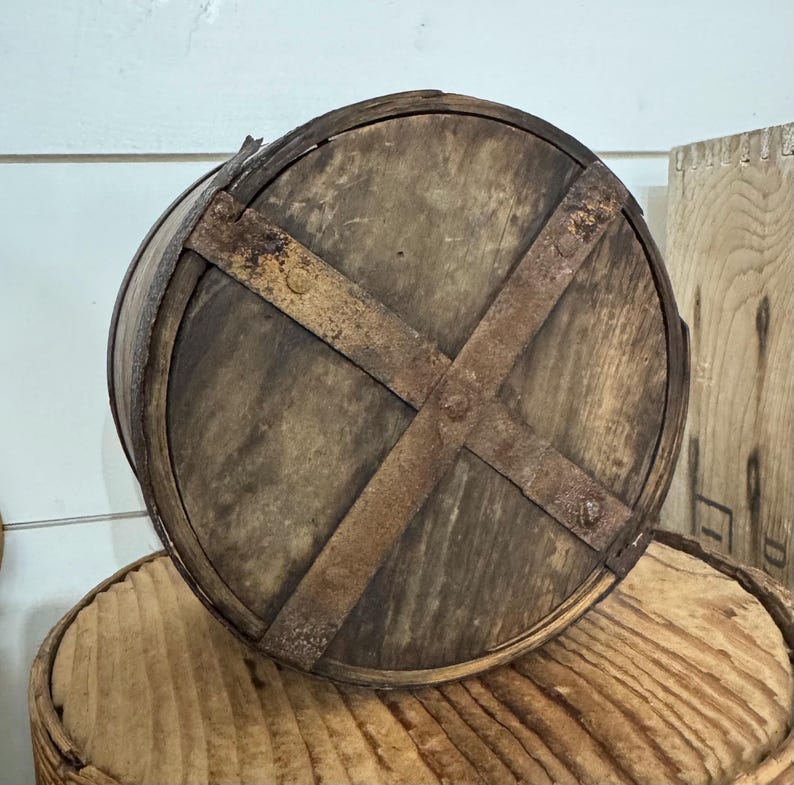May include: A rustic, wooden barrel with a circular base and a weathered appearance. The barrel is reinforced with metal bands that cross in an X-shape, adding to its vintage aesthetic. The wood has a natural, aged color.