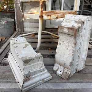 May include: Two architectural corbels featuring a distressed white painted finish over wood. Each corbel displays a curved profile with visible chipped paint, revealing the natural wood texture underneath, and includes pre-drilled mounting holes.