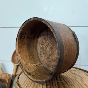 May include: A vintage wooden measuring cup with a dark brown metal band. The interior features stamped text, including "DANIEL'S MANUFACTURE" and "WILT N". The cup is cylindrical and shows signs of age and wear.