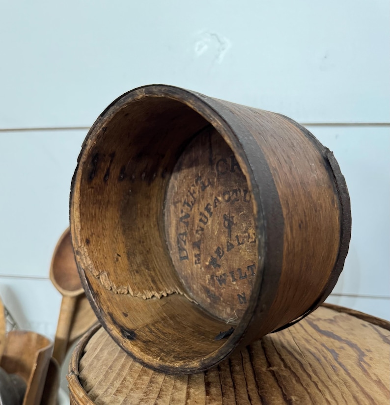 May include: A vintage wooden measuring cup with a dark brown metal band. The interior features stamped text, including "DANIEL'S MANUFACTURE" and "WILT N". The cup is cylindrical and shows signs of age and wear.