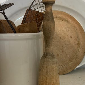 May include: A vintage wooden muddler with a rounded top and a flared base stands in front of a white ceramic container. Other antique kitchen tools, including a wooden cutting board and a wire whisk, are visible.